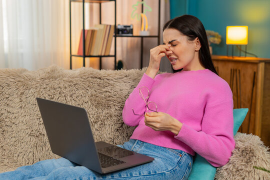 Young woman at home closing laptop after prolonged work, removing glasses and rubbing tired eyes. Caucasian girl on sofa experiencing fatigue, exhausted from long screen time, needing rest break.