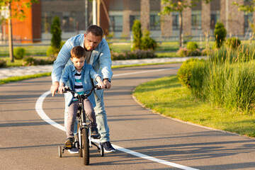 Father teaching son riding bike on the bike path. Father helping excited son to ride a bicycle in modern urban district. Child learning to ride cycle with his dad. Fathers day.