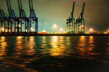 Nighttime View Of Cargo Cranes  in the harbor at night, illuminated by bright lights against a dark sky. The calm surface of the water reflects the bright colors in a nighttime atmosphere.
