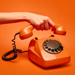 Hand lifts the receiver of a vintage orange rotary dial telephone against a matching orange...