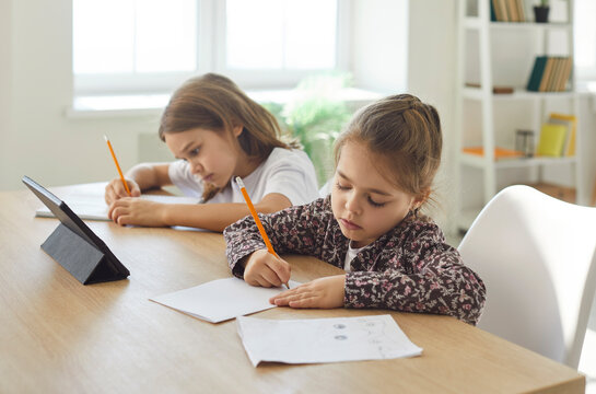 Two children of different age sitting at table together, writing with pencils and doing homework. Focused siblings studying or drawing, using tablet for learning. Homeschooling and leisure concept.