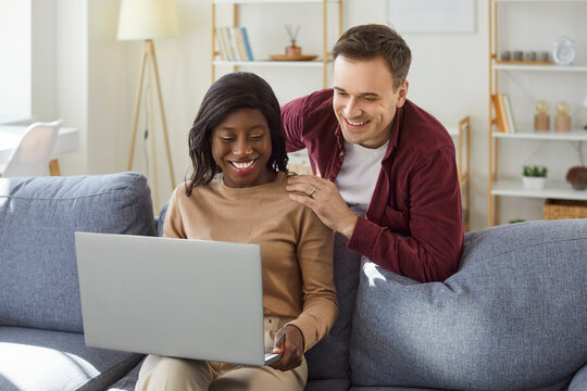 Young happy couple sitting on sofa in living room at home together watching funny video or using laptop for internet and social media. Happy man and African American woman resting on couch.