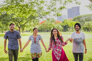 Group of four Indian friends walking in a park, smiling and holding hands. Diverse group enjoying a sunny day outdoors, surrounded by trees and greenery. Young Indian friends at the park.