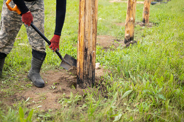 Worker in protective gloves using a shovel install a wooden post for a fence. Manual labor, land management, environmental conservation, or DIY construction projects.