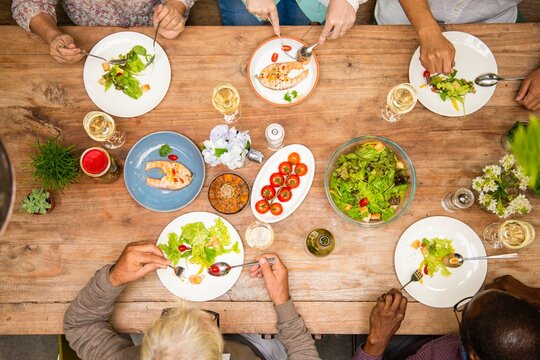 Top view of diverse group enjoying a meal. Plates with salad, chicken, and tomatoes on a wooden table. Hands reaching for food, sharing a communal dining experience. Lunch food on dinner table.