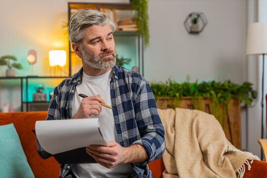 Strict bearded mature man in casual clothes with documents analyzing information reading business papers prepare financial report. Senior Caucasian old guy shocked by sudden victory success on sofa.