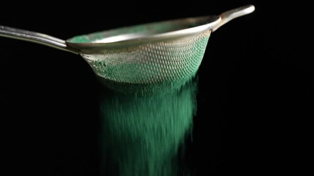 Sifting natural green spirulina powder through a metal sieve on black background, slow motion. Close up organic spirulina algae powder. Dietary supplement to improve health. Seaweed superfood concept