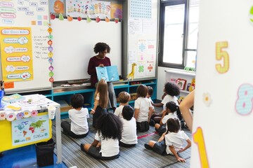 Teacher reading to diverse group of children in classroom. Kids sitting on floor, listening. Classroom setting with books, posters, and educational materials. Education and knowledge concept.