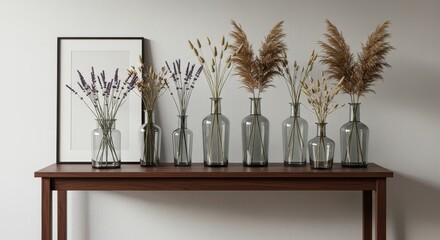 Naklejka premium Dried Flowers in Glass Vases on Console Table - Seven glass vases of varying heights hold dried lavender and pampas grass, arranged on a dark brown console table against a white wall