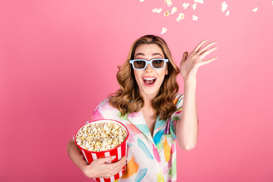 Cheerful young woman with popcorn tossing kernels playfully against pink background in colorful attire, enjoying fun leisure time.