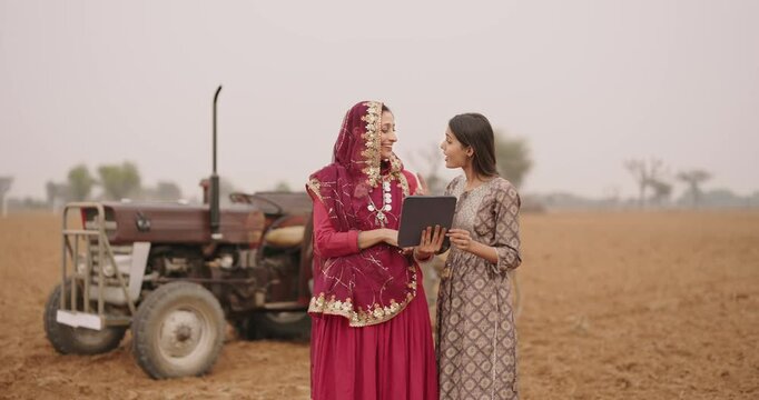 Indian asian villager farmer young adult woman lady wear red saree smiling outdoor hold tab device browse website data happy teen age girl teach to mom talking gossip enjoy day time outside desi life