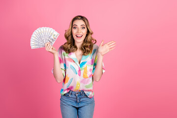 Enthusiastic young woman holding cash and waving her hand against a cheerful pink background,...