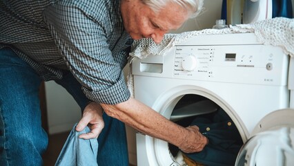 Elderly man doing laundry, placing clothes in washing machine. Senior male, casual attire, home setting. Laundry, washing, clothes, home chores. Senior man doing laundry chores at home.