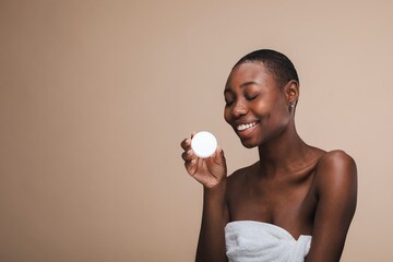 Smiling person holding a skincare product, wrapped in a towel, against a neutral background. Focus...