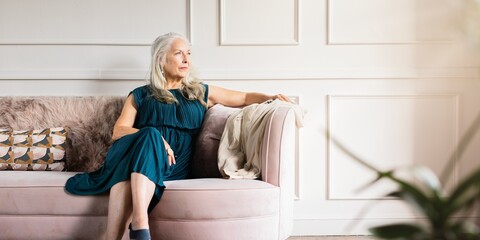 Elegant older woman wearing dress, sits on couch. The older woman looks thoughtful. Elegace old...