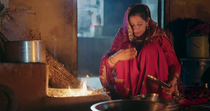 Indian villager woman preparing food using traditional earthen mud stove indoor house asian young female wear red saree cooking on open fire flame inside kitchen home