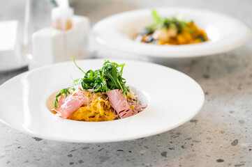  A beautifully plated pasta dish with ham and fresh arugula sits in the foreground on a textured surface, while a second plate of pasta is blurred in the background, showcasing culinary elegance.
