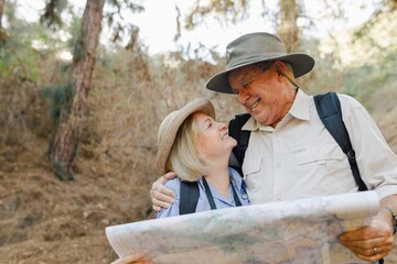 Elderly couple hiking with a map. Smiling elderly man and woman hiking together. Elderly hikers enjoying nature. Elderly couple exploring outdoors. Elderly man holding map and embrace his wife