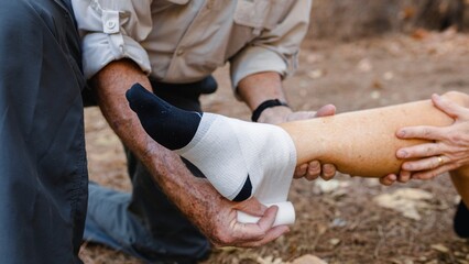 Person bandages another's ankle outdoors. People doing first aid to  another's ankle while hiking outdoor. Man help first aid and bandages injured ankle in natural outdoor. Injured ankle hiking