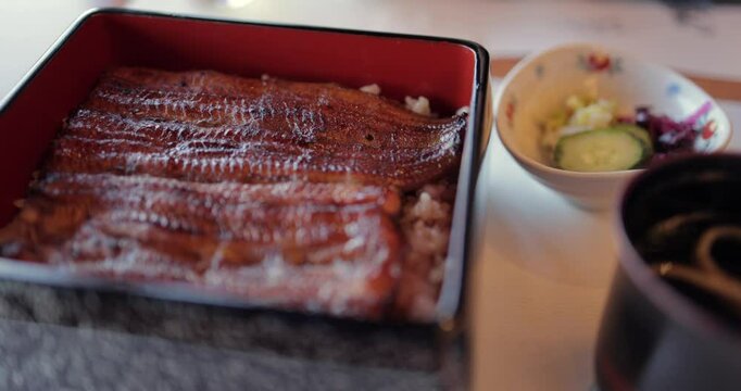 Close-up shot of traditional Japanese unagi donburi with glazed grilled eel over rice in a lacquered box as the camera makes a slow semicircular movement above the dish