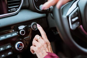 Fototapeta premium Close-up of a hand pressing a car's start button. The hand is adorned with rings, and the dashboard is visible. The start button is prominently featured. Senior woman turning on car.