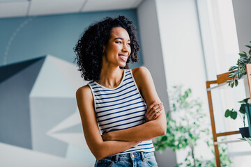 Young woman with curly hair smiling confidently indoors on a sunny day, embracing a comfortable and...