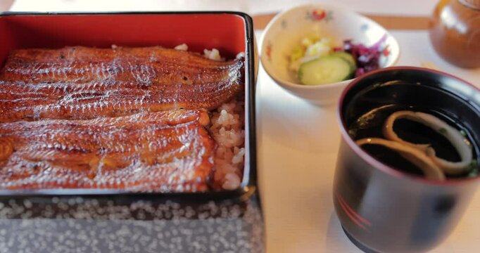 Smooth lateral camera movement reveals a complete Japanese meal set with grilled eel donburi, miso soup, and fermented sides served in traditional lacquerware from above