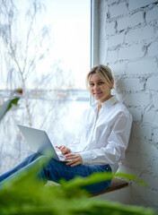 A young woman in a white button-down shirt and blue jeans sits by the window using a laptop. The girl is smiling, freelancing, business, teaching. Behind her back is a window and to her right is a