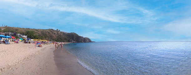 A panoramic view of a crowded sandy beach with colorful umbrellas and people enjoying the sunny weather next to a clear blue sea and a rocky coastline. Capo Vaticano, Calabria, Italy