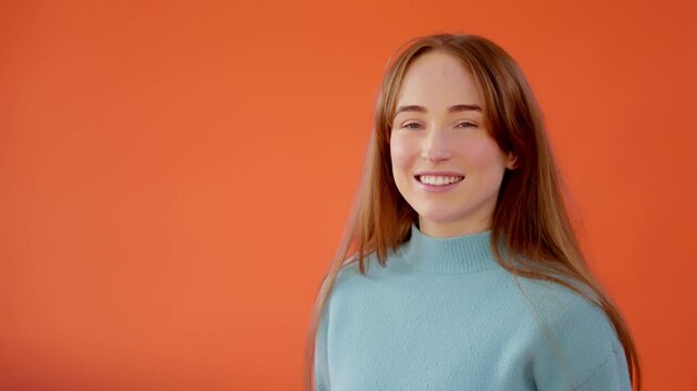 Young woman with long brown hair smiles warmly against vibrant orange background. She wears  light blue sweater, creating cheerful and inviting atmosphere