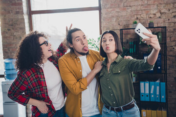 Three cheerful young coworkers having fun taking a selfie together in a modern casual office...