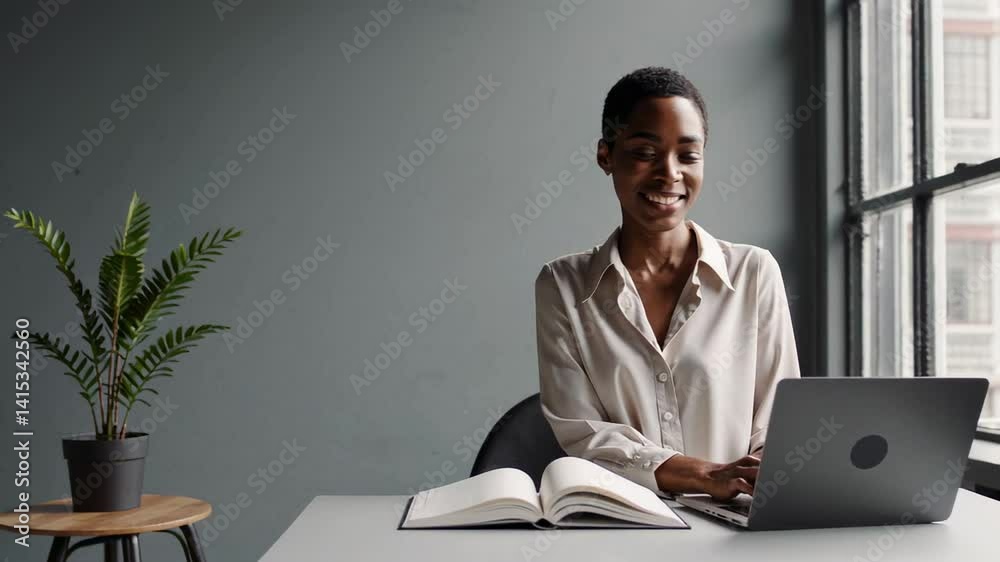 Poster office worker smiles while typing on her laptop with a notebook open in front of her, near a window 