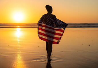 woman standing on beach at sunset wrapped in american flag silhouette