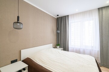cozy bedroom featuring a neatly made bed, modern pendant lights, and window with sheer and gray curtains. Neutral tones create a calming atmosphere