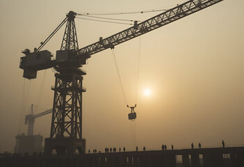 Towering Crane Lifting Steel Beams at Sunrise &ndash; Silhouetted Workers Amidst Foggy Dawn