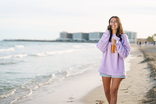 Happy Mediterranean Woman In Oversized Lavender Sweater Strolling Barefoot On Sandy Beach With Cold Beer - Powered by Adobe