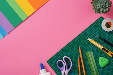 Supplies for paper art hobby: scissors, craft knife, pencil, eraser, cutting mat, colored paper on pink background. Copyspace. Paper crafts work table flatlay.