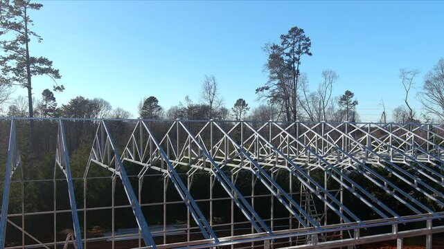 Metal framework of a large greenhouse under construction with triangular roof trusses and vertical supports. The structure features a series of parallel steel beams creating the skeleton of what will