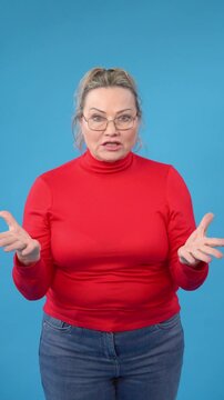 Older woman in  red turtleneck sweater expressing surprise or confusion against  blue background. She has glasses and is gesturing with  hands, conveying  sense of disbelief or questioning