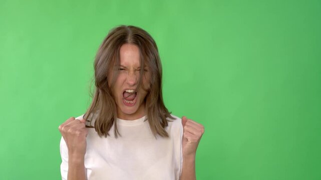 Woman with long brown hair is expressing intense emotion, with her fists clenched and mouth open in  yell. She is wearing a plain white t-shirt and is set against  bright green background.