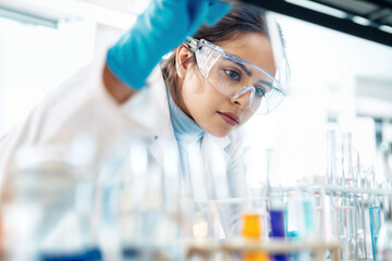 The image shows a scientist in a laboratory setting, wearing a white lab coat and blue gloves, inspecting a rack of test tubes containing colorful liquids