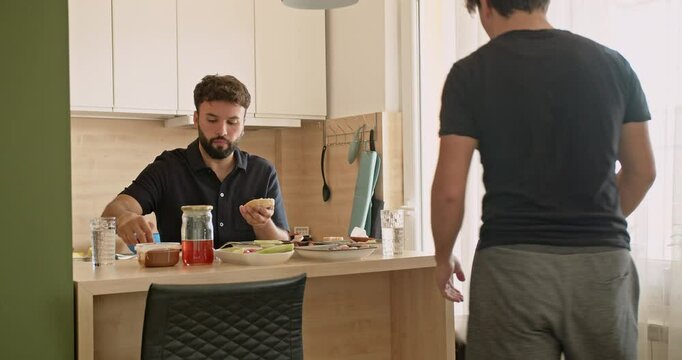 Two Caucasian men are having breakfast in a modern kitchen. One man is spreading butter on a slice of bread while the other sits down to join him.