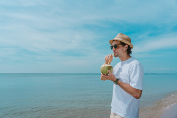 Tourist having a coconut drink on the beach. Young handsome man drinking coconut milk on the tropical beach, relaxing. Summer vibes. Smiling man holding fresh coconut drink by the shore on the beach