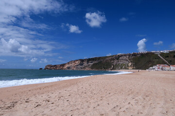 A sunny beach scene with golden sand, gentle waves and a rocky cliff topped with houses under a vibrant blue sky in Nazaré, Portugal.