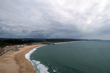 Fototapeta premium Panoramic view of a curved beach flanked by a coastal town and backed by green hills, under a cloudy sky stretching out over the ocean, in Nazaré, Portugal. 
