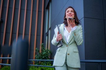 Smiling businesswoman wearing suit holding folder and recording voice message on smartphone while...