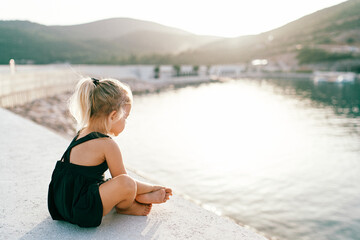 Little barefoot girl sits cross-legged on the pier and looks at the water. Side view