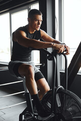 Handsome athletic man in sportswear is working hard on an air bike, performing a cardio workout in a modern gym. His focus and intensity show his commitment to a healthy lifestyle and fitness goals.