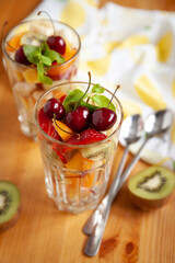 Directly above on large transparent glasses with chopped fruits and berries and long spoons on the table. Tangerine, kiwi, banana, strawberries, mango, cherries and mint leaves. Healthy snack.