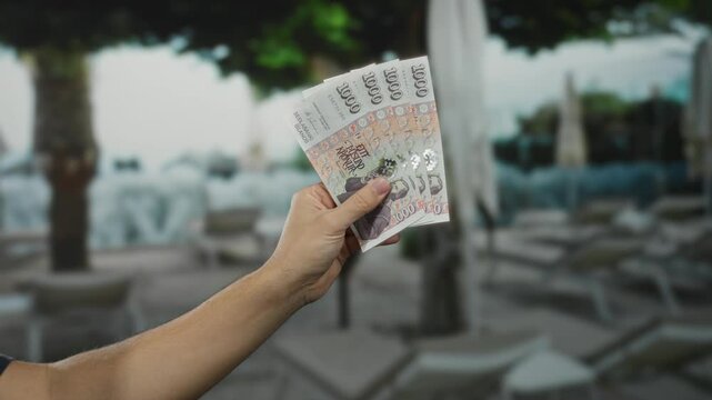 Male hand holding icelandic krona banknotes at outdoor hotel pool with trees and lounge chairs visible in the background suggesting vacation spending.